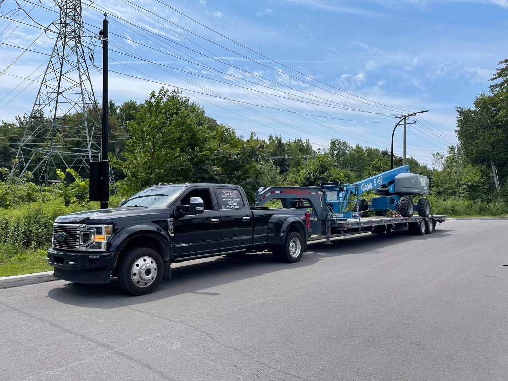 Camionnette noire Transport JKM et nacelle bleue sur remorque.