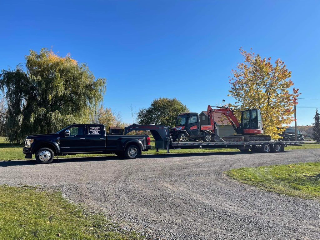 Camionnette Transport JKM et remorque portant une excavatrice rouge et un petit véhicule.