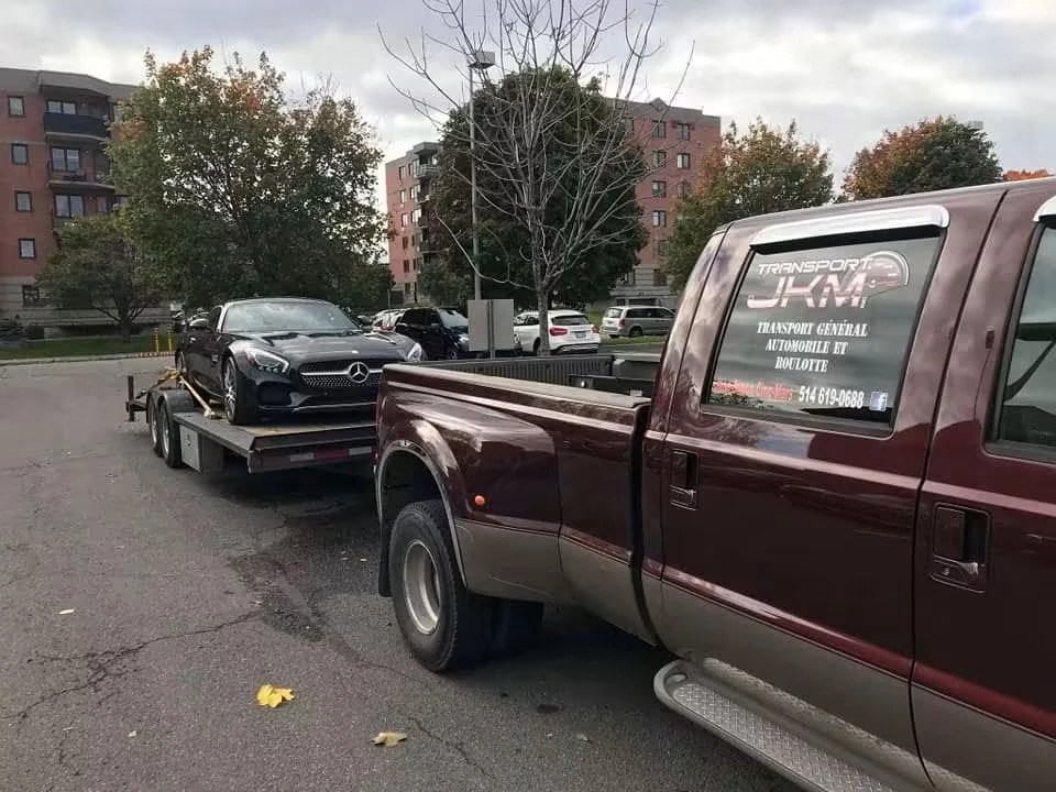 Mercedes noire sur une remorque à plateforme ouverte, derrière une camionnette Transport JKM.