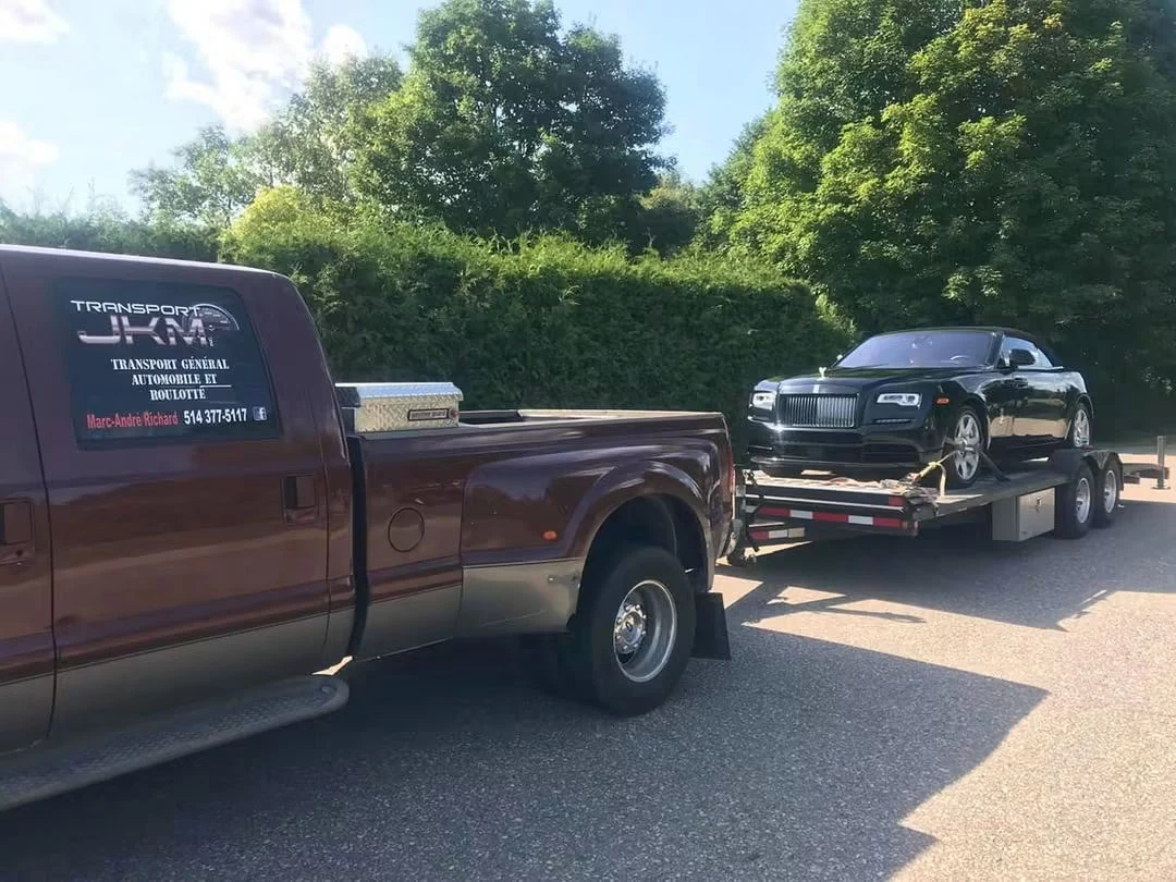 Black Rolls-Royce on an open flatbed trailer behind a Transport JKM pickup truck.